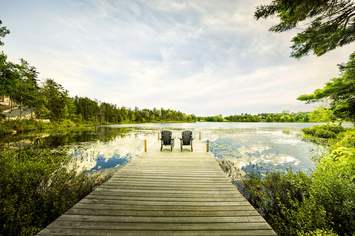 image shows two chairs at the end of a dock overlooking a lake surrounded by trees with a blue sky