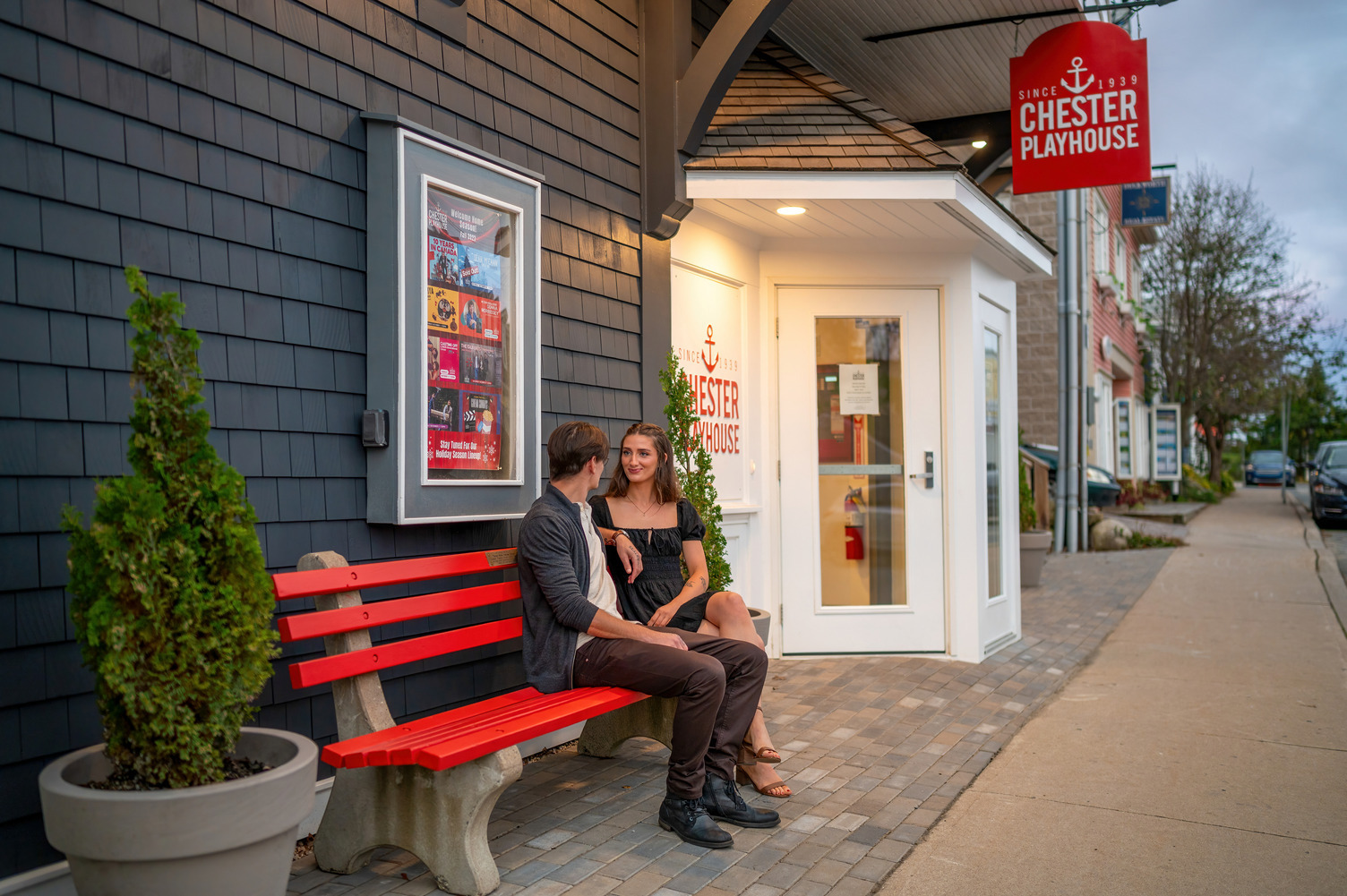 Two people sitting on a red bench outside the chester playhouse in the early evening.